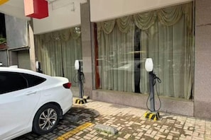 A car parked outside a building with two charging stations, a tiled pavement, and a window with curtains. - Guangyuezhen Hotel (Baise High-Speed Railway Station) (Baise)