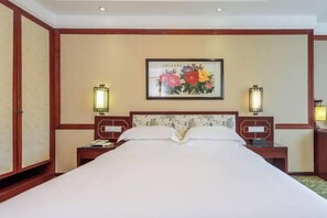 Una habitación de hotel con una cama grande, dos mesitas de noche, una lámpara de noche y una obra de arte floral enmarcada en la pared, además de dos lámparas fijadas a la pared.