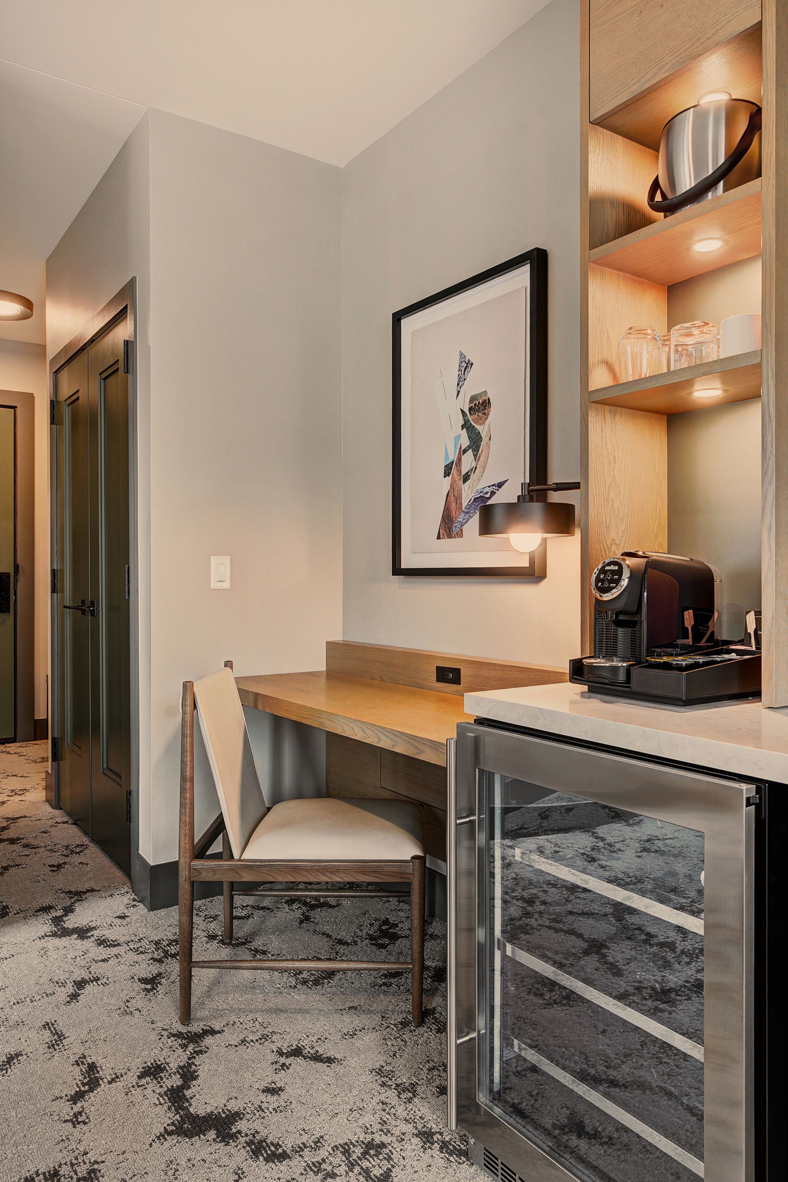 Une chambre d’hôtel  équipée d’un bureau, d’une chaise, d’une machine à café, d’œuvres d’art encadrées et d’une étagère contenant des ustensiles de cuisine.