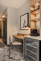 A hotel room with a desk, chair, coffee machine, framed artwork, and a shelf with kitchenware.