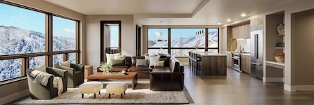 A modern living room with a sofa, coffee table, and ottoman, a kitchen with a refrigerator and oven, and large windows with a snowy mountain view.