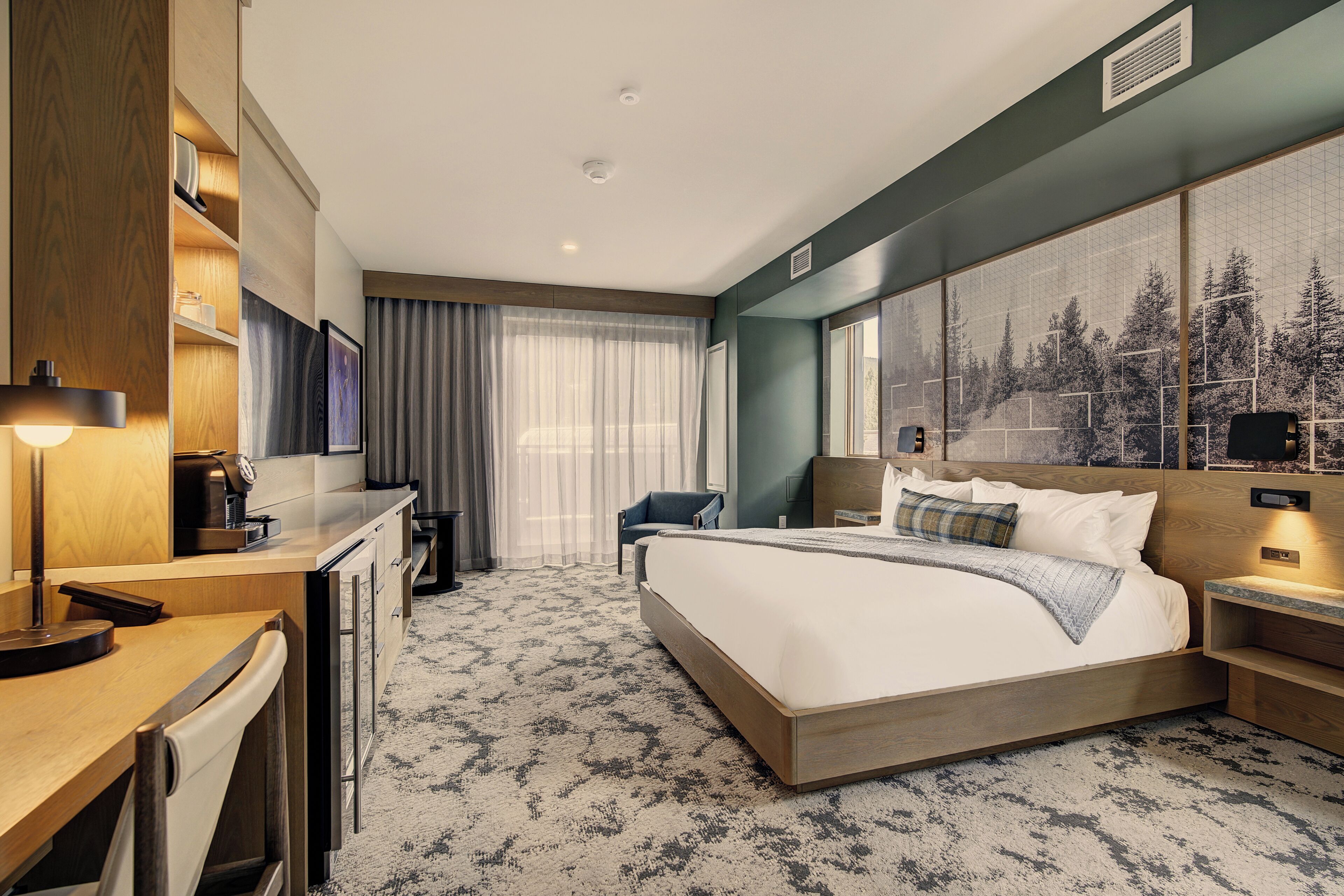 Ein Hotelzimmer mit einem großen Bett, einem Schreibtisch mit Lampe, einem Sessel, einem Fernseher und einem großen Fenster mit Blick auf einen Wald.