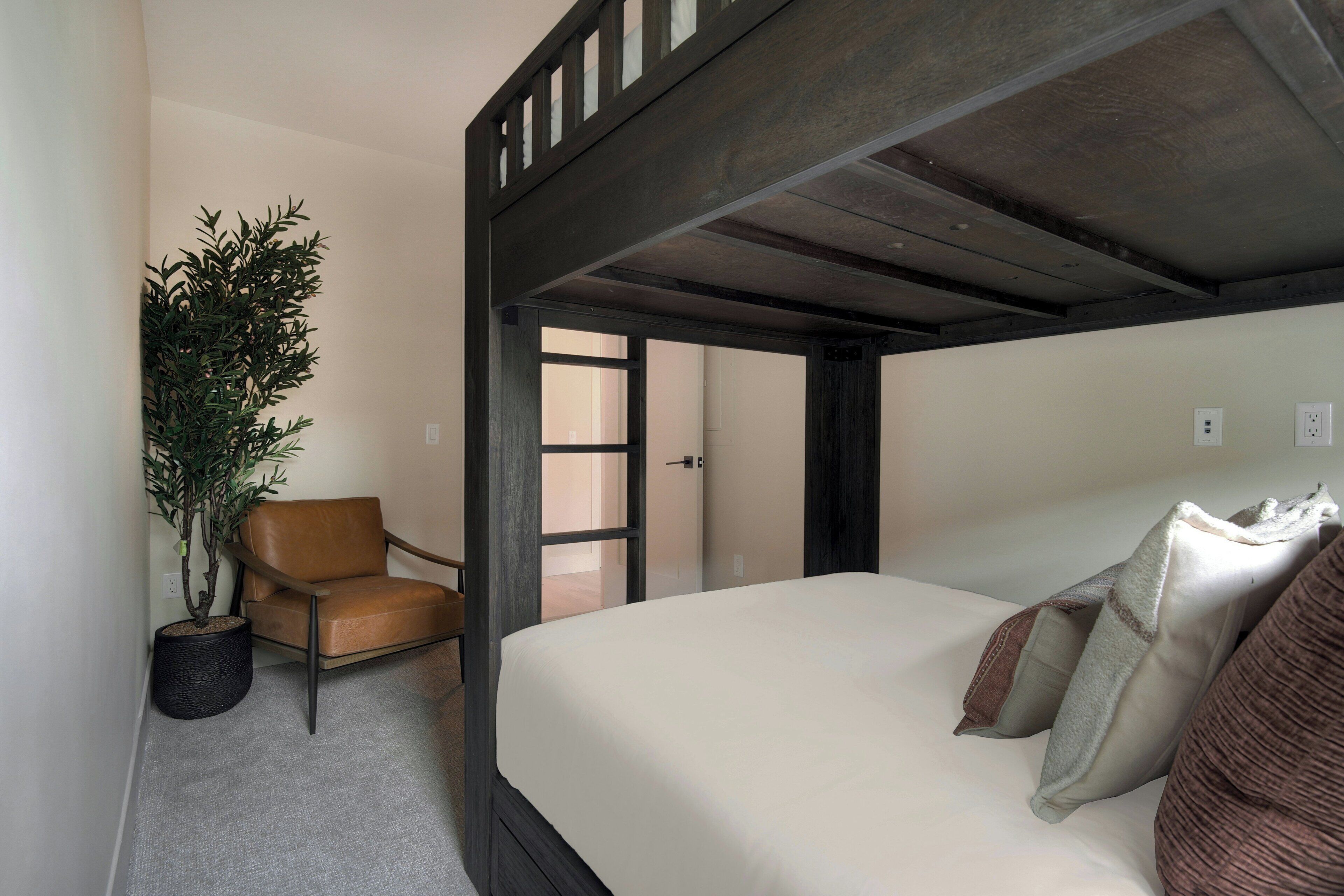 A bedroom with a four-poster bed, a potted plant, a leather chair, and a door leading to another room.