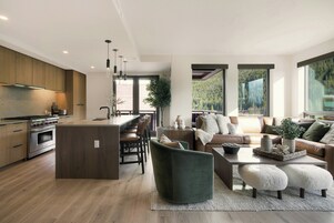 Una cocina moderna con una isla central, comedor con sillas, un conjunto de sofás y ventanas amplias con vistas a un bosque.