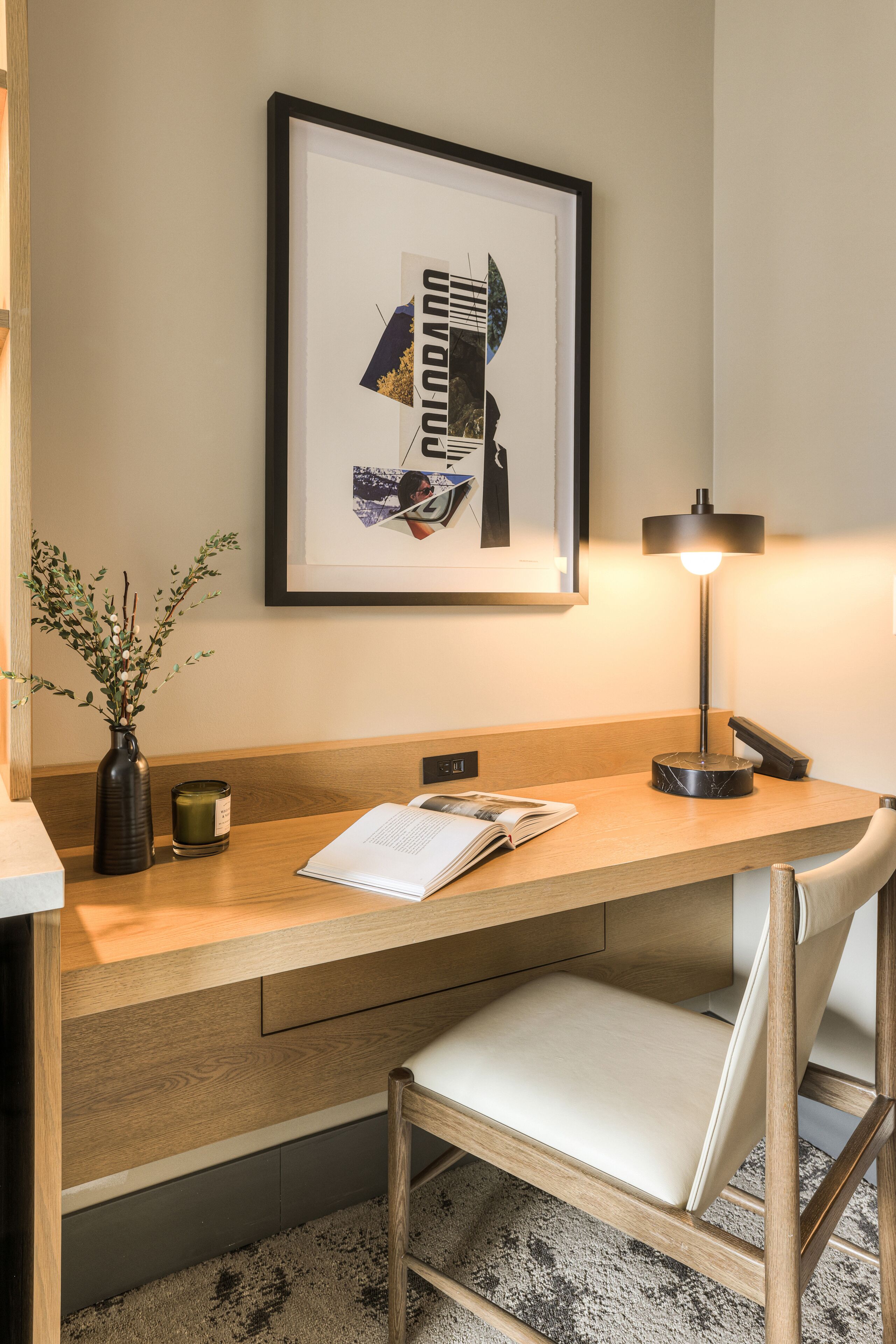 A hotel room with a wooden desk, a framed artwork, a vase with greenery, a candle, and a lamp.