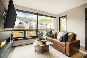 A modern living room with a leather sofa, a flat-screen TV mounted on the wall, and a large window offering a view of neighboring buildings and a mountain. - Kindred Resort a RockResort (Keystone)