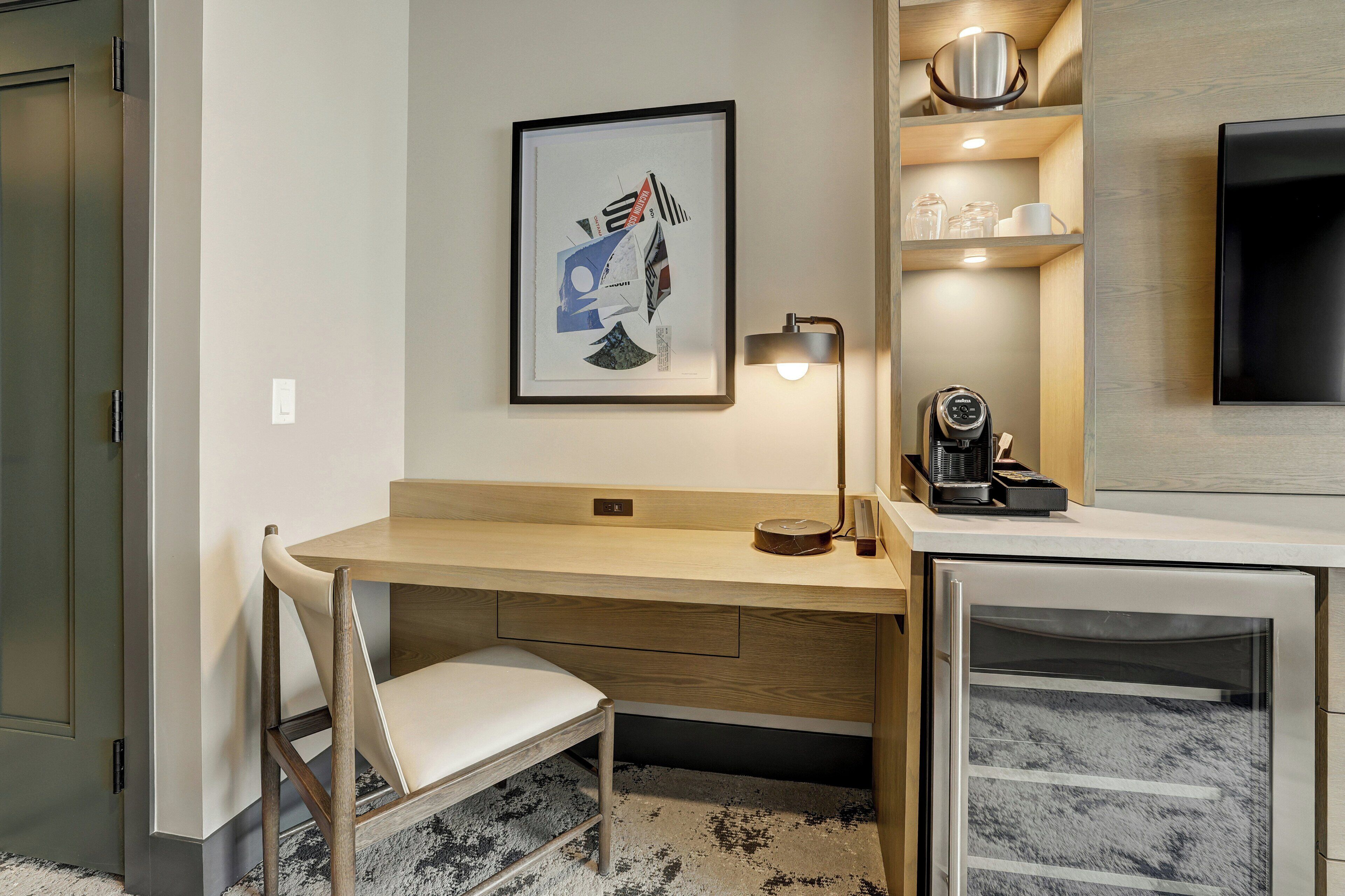 Una habitación de hotel moderna con un escritorio de madera, una silla, una obra de arte enmarcada, una cafetera y un televisor.