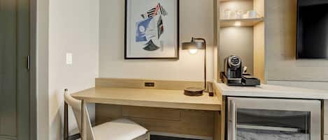 A modern hotel room with a wooden desk, a chair, a framed artwork, a coffee machine, and a television.