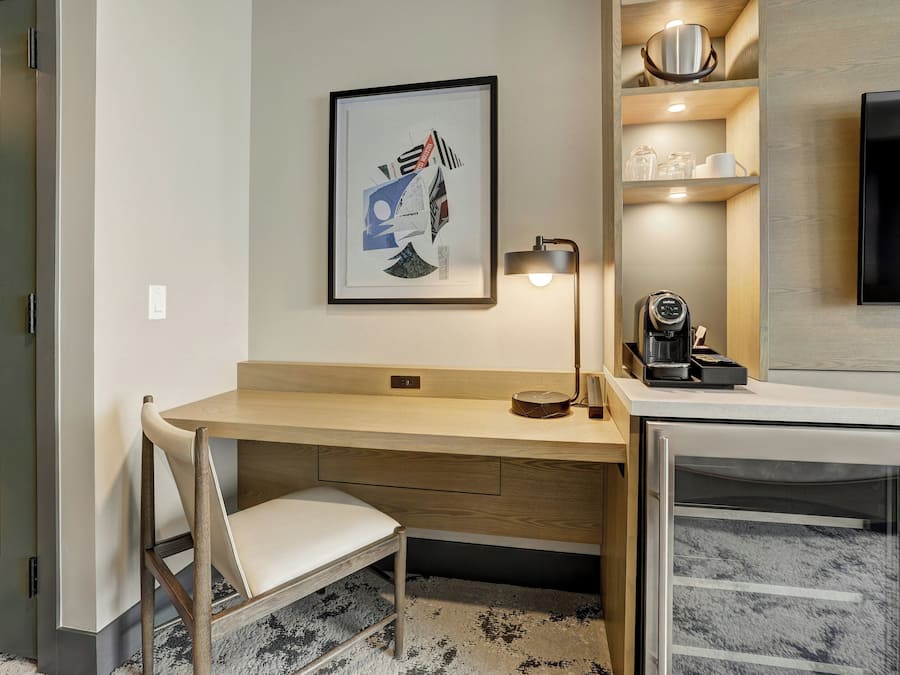 A modern hotel room with a wooden desk, a chair, a framed artwork, a coffee machine, and a television.