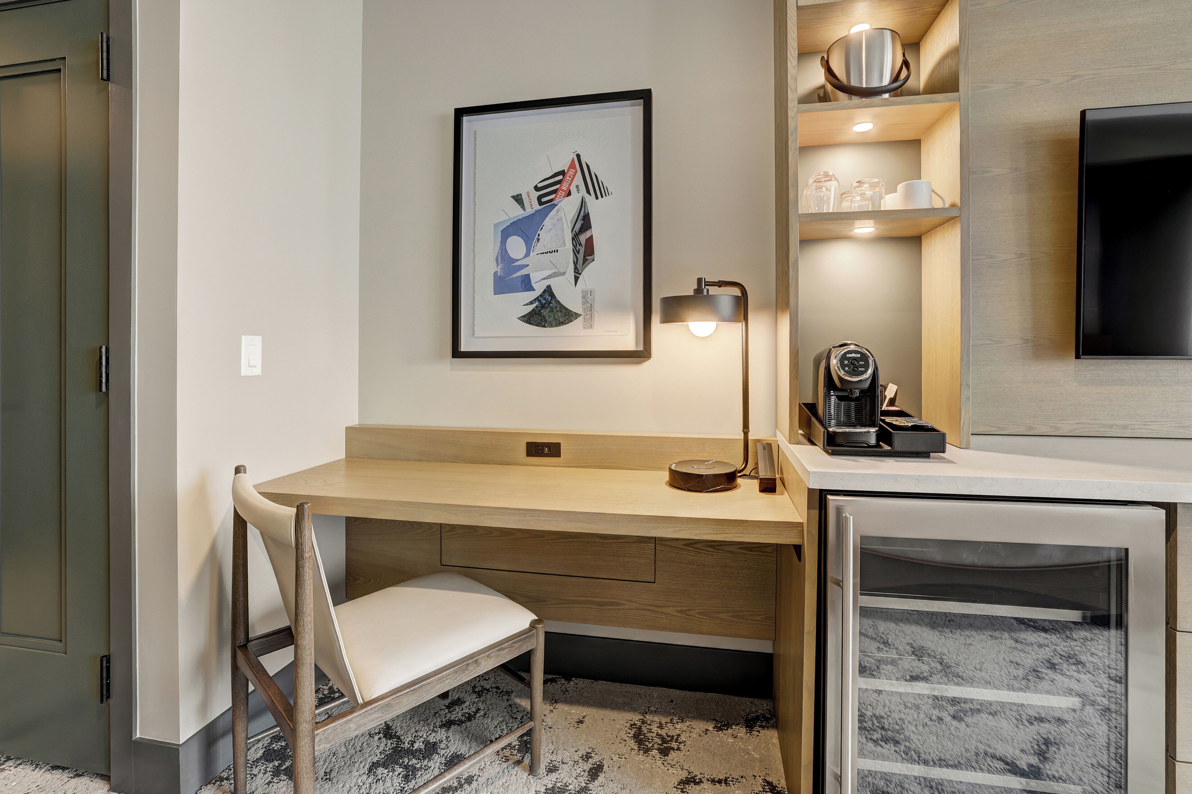 A modern hotel room with a wooden desk, a chair, a framed artwork, a coffee machine, and a television on the wall.