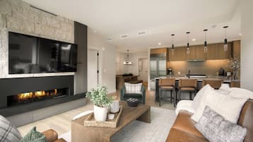 A modern living room with a fireplace, a flat-screen TV, and a kitchen area with wooden cabinets and pendant lighting.