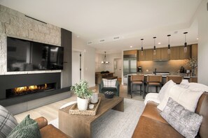 A modern living room with a fireplace, a flat-screen TV, and a kitchen area with wooden cabinets and pendant lighting.