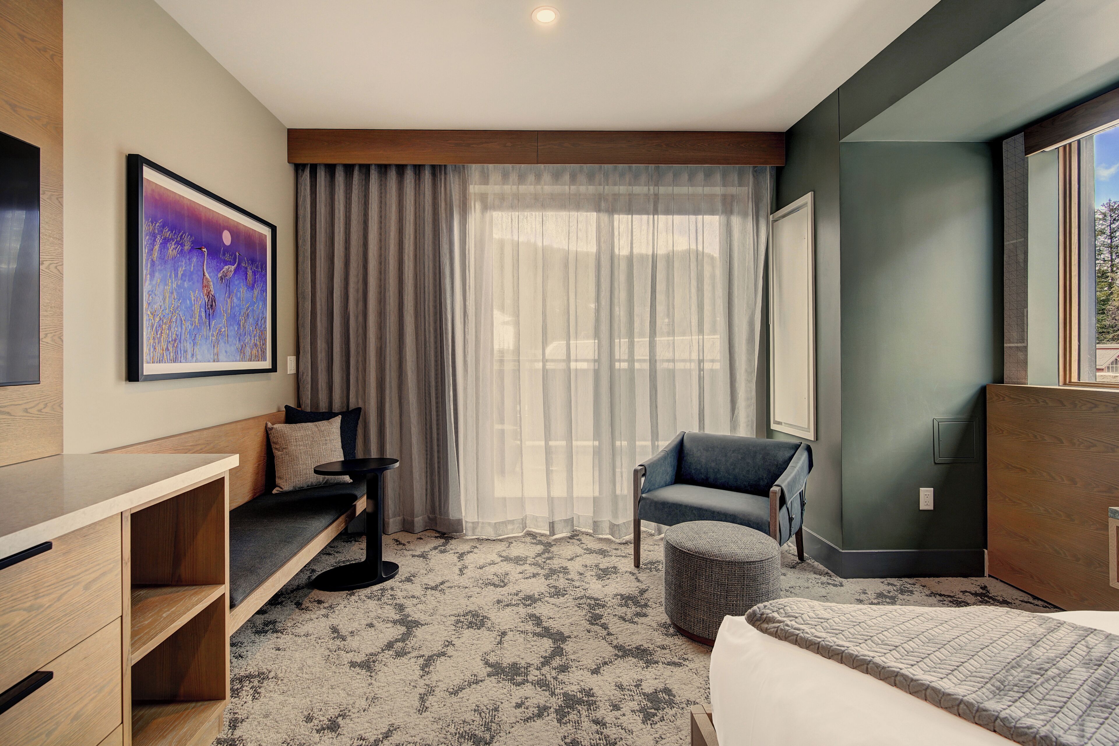 A hotel room with a bed, a sofa, a chair, a small table, and a large window with curtains.