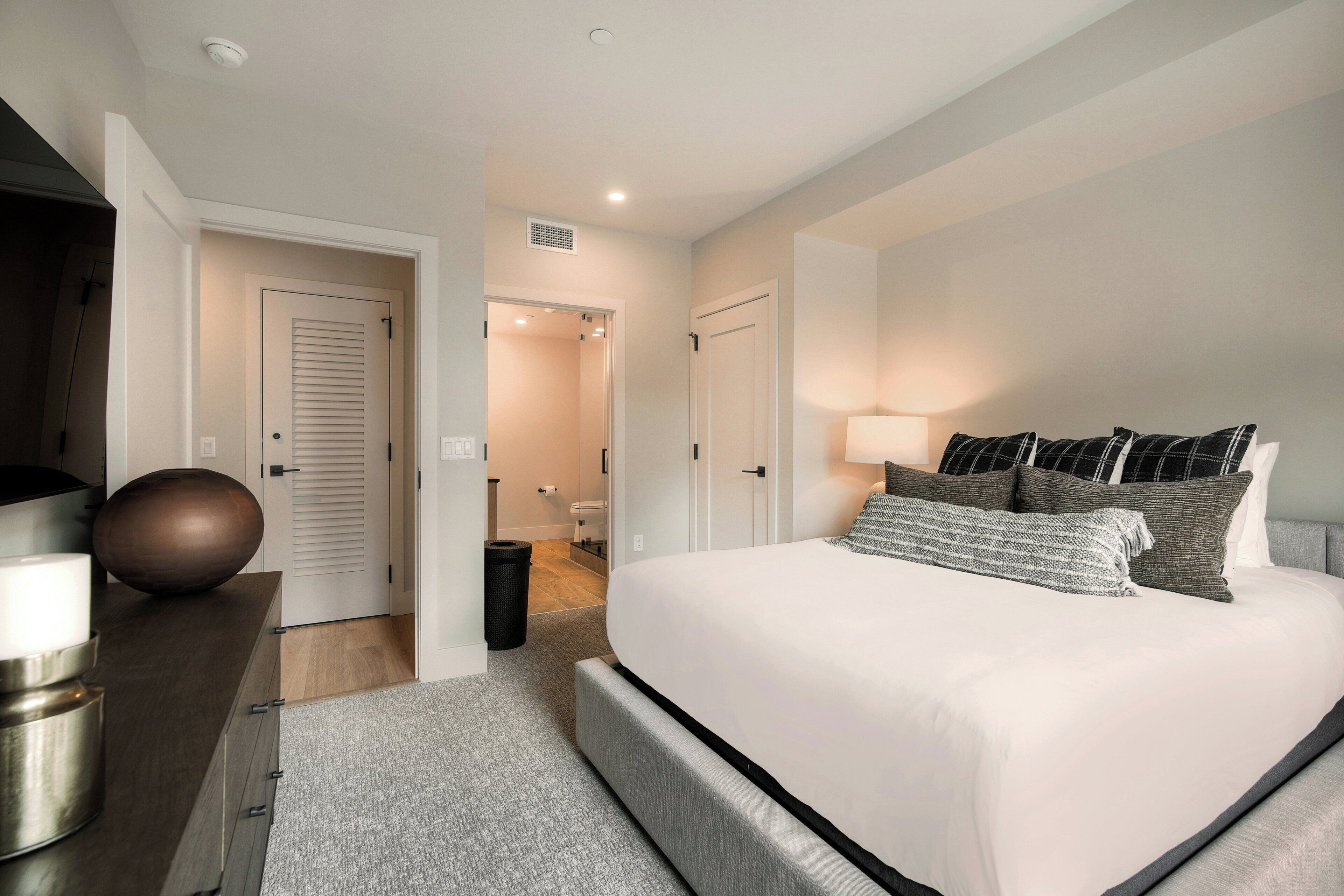 A modern bedroom with a large bed, bedside tables, a lamp, and a view into the bathroom.