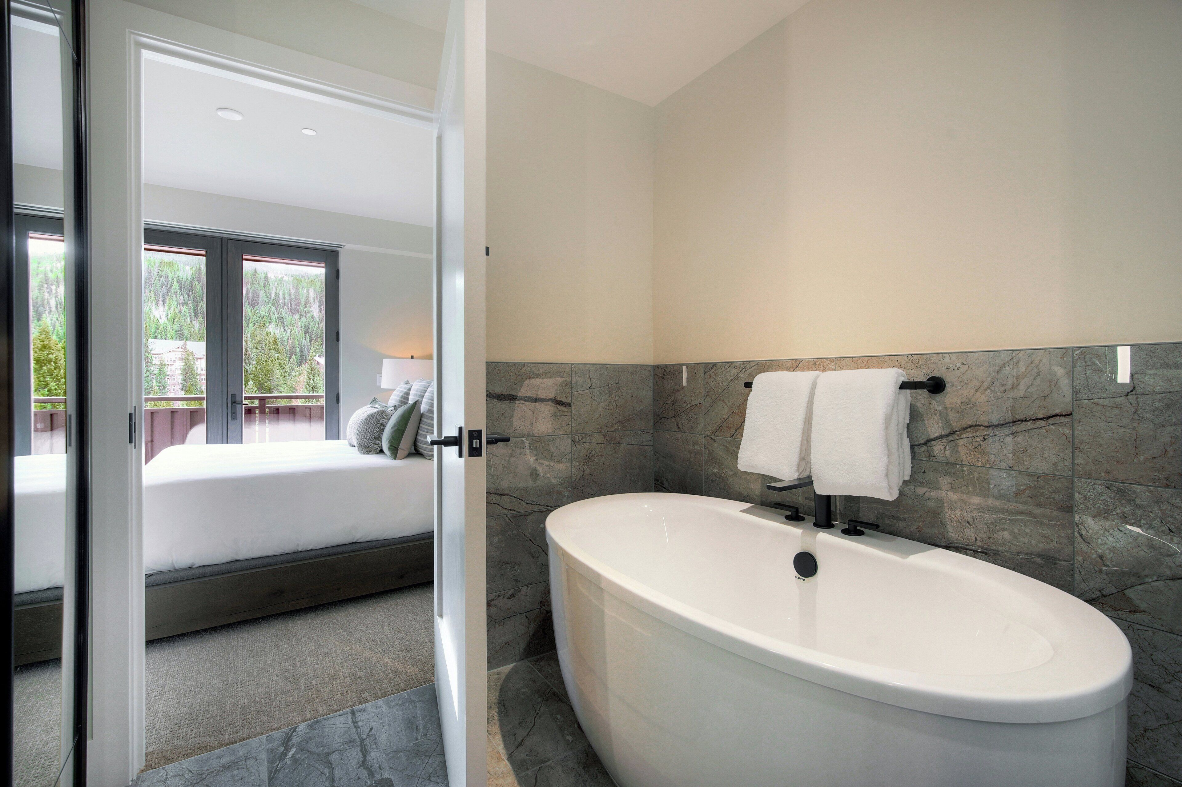 A modern bathroom with a freestanding bathtub, a towel rack, and a view of a bedroom with a bed and a window.
