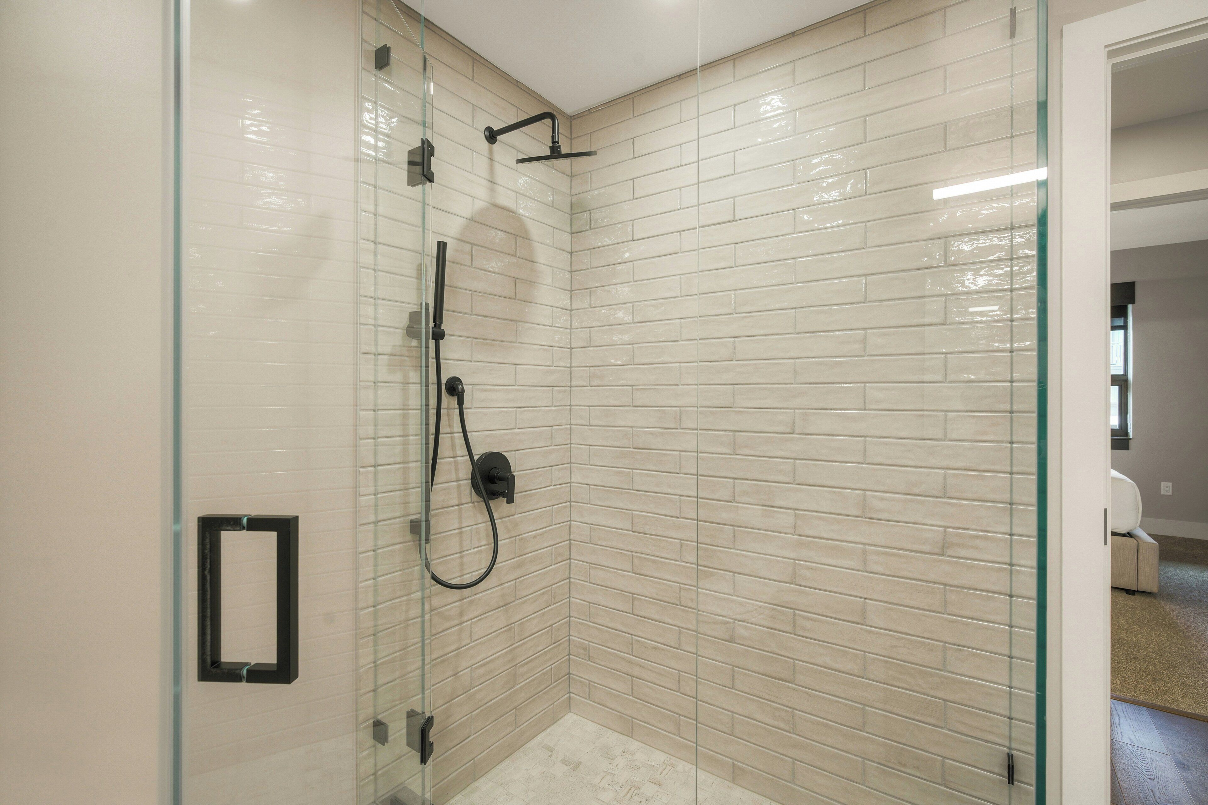 A glass-enclosed shower with a tiled wall, a visible shower control panel, and a door leading to another room.
