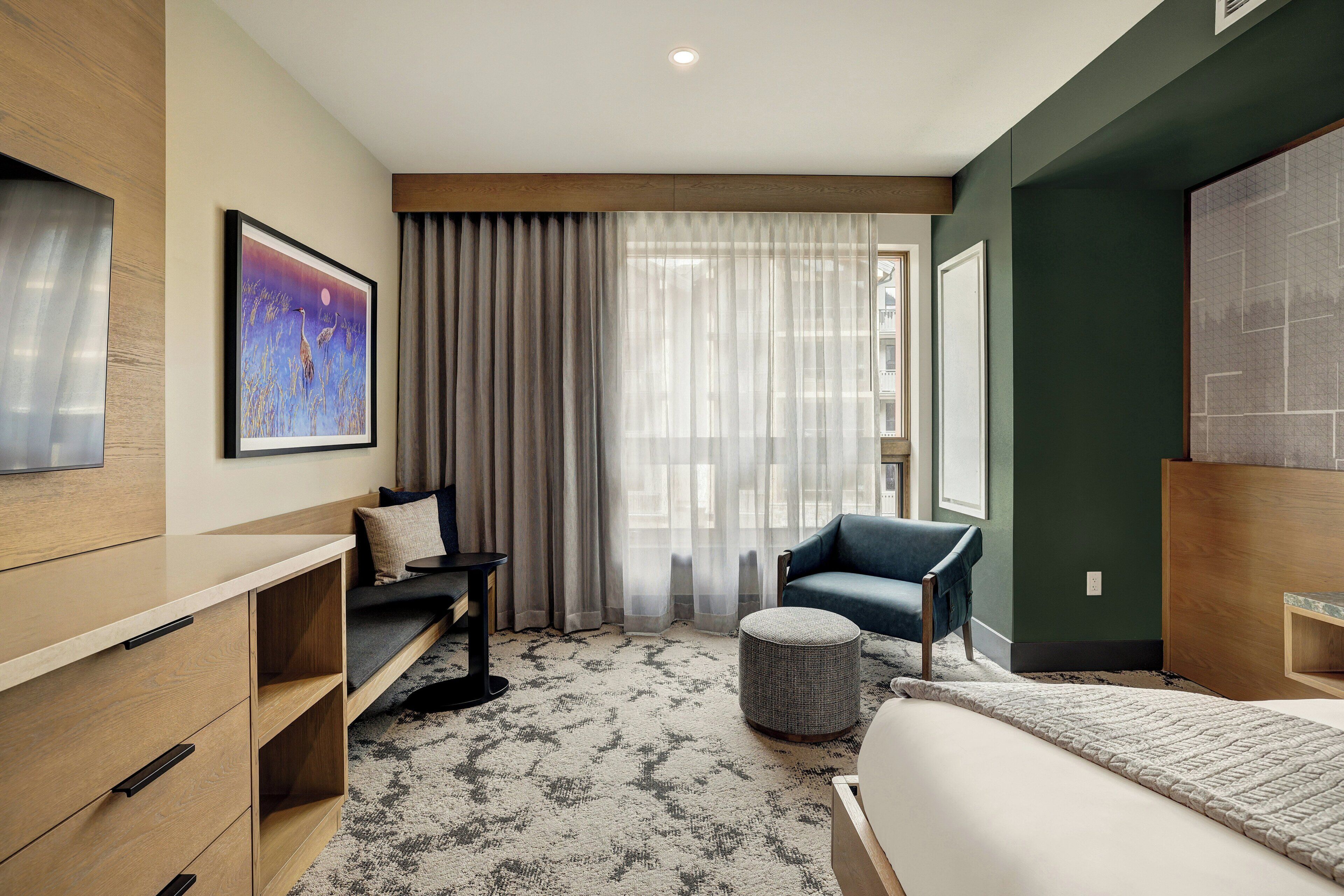 A modern hotel room with a bed, a sofa, a small table, and a chair. There is a painting on the wall and a window with curtains.