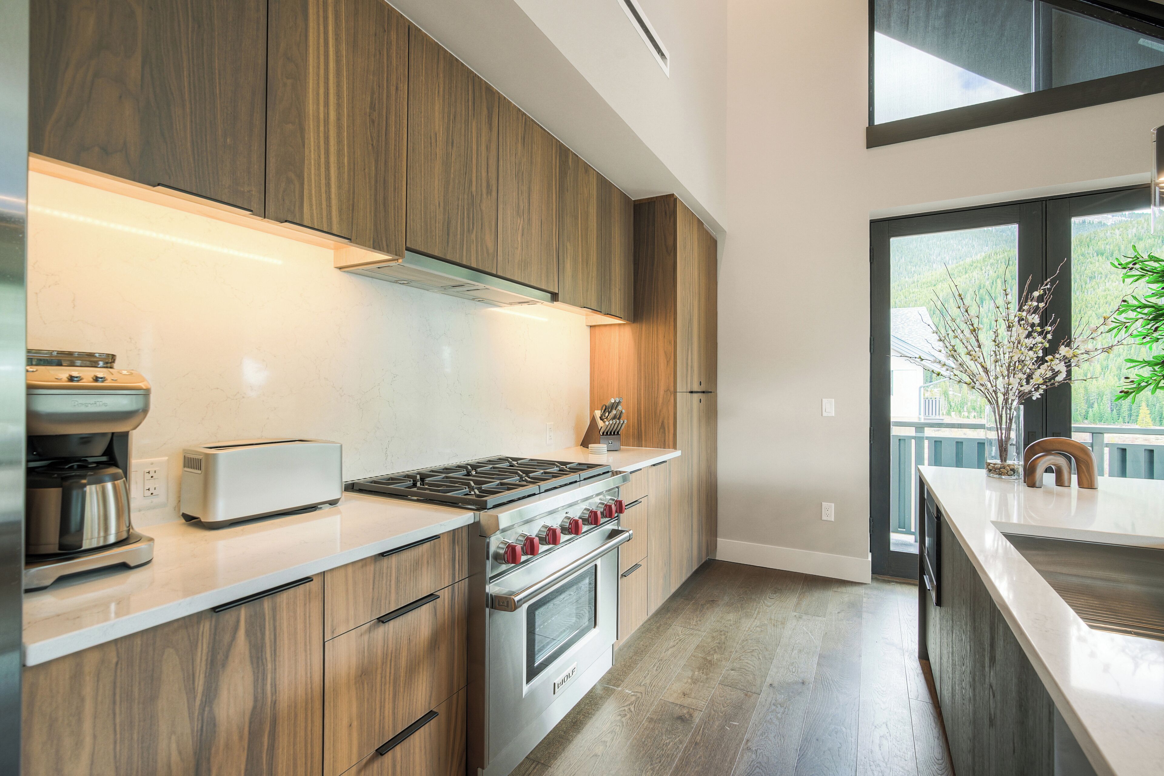 A modern kitchen with wooden cabinets, stainless steel appliances, and a view of a green landscape through the window.