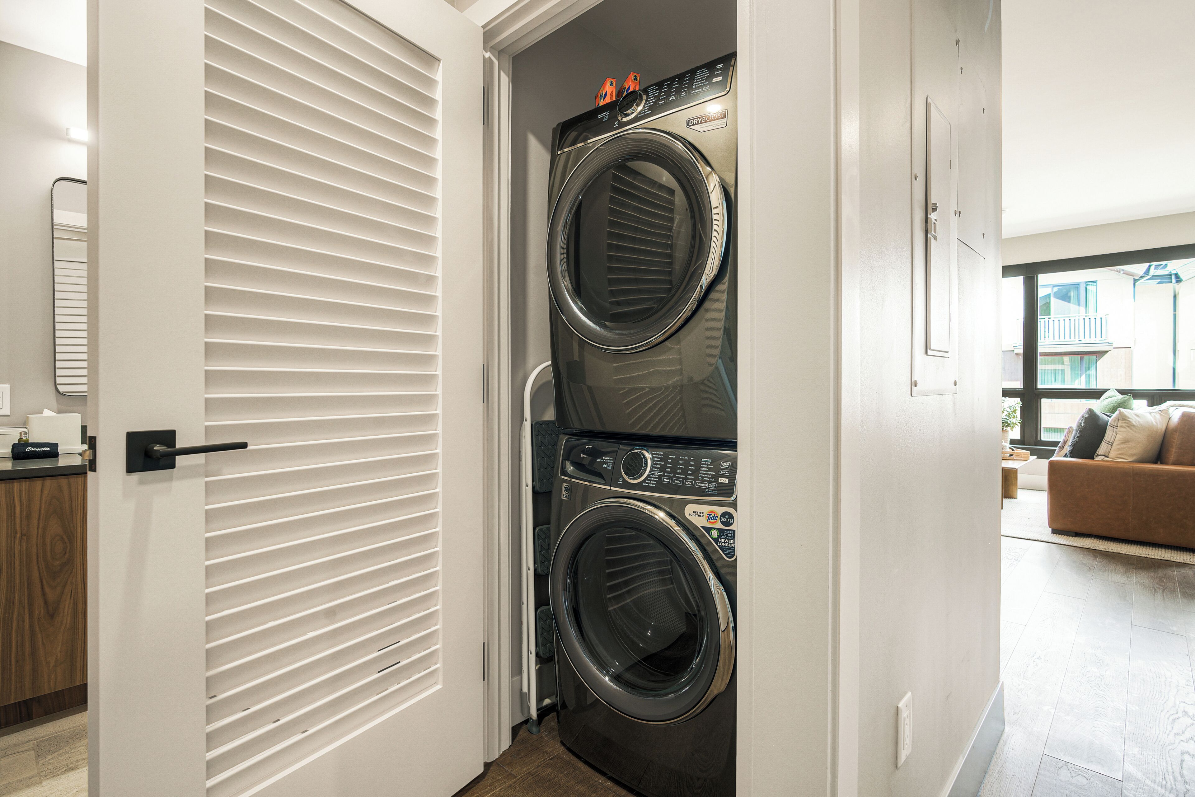 A laundry room with two stacked front-loading washers and dryers, a door with horizontal blinds, and a view of a living area with a couch and a window.