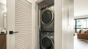 Un lavadero con dos lavadoras y secadoras apiladas, una puerta con persianas horizontales y vista a una sala de estar con sofá y ventana.