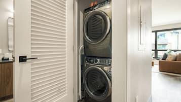 A laundry room with two stacked front-loading washers and dryers, a door with horizontal blinds, and a view of a living area with a couch and a window.