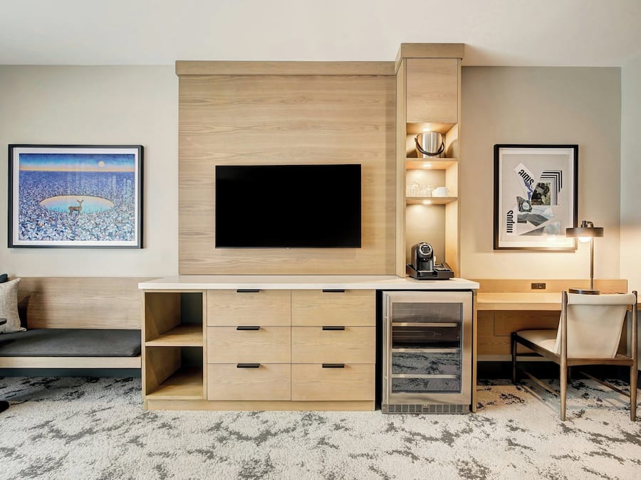 A modern hotel room with a flat-screen TV mounted on a wooden wall, a desk with drawers, a chair, and a sofa.