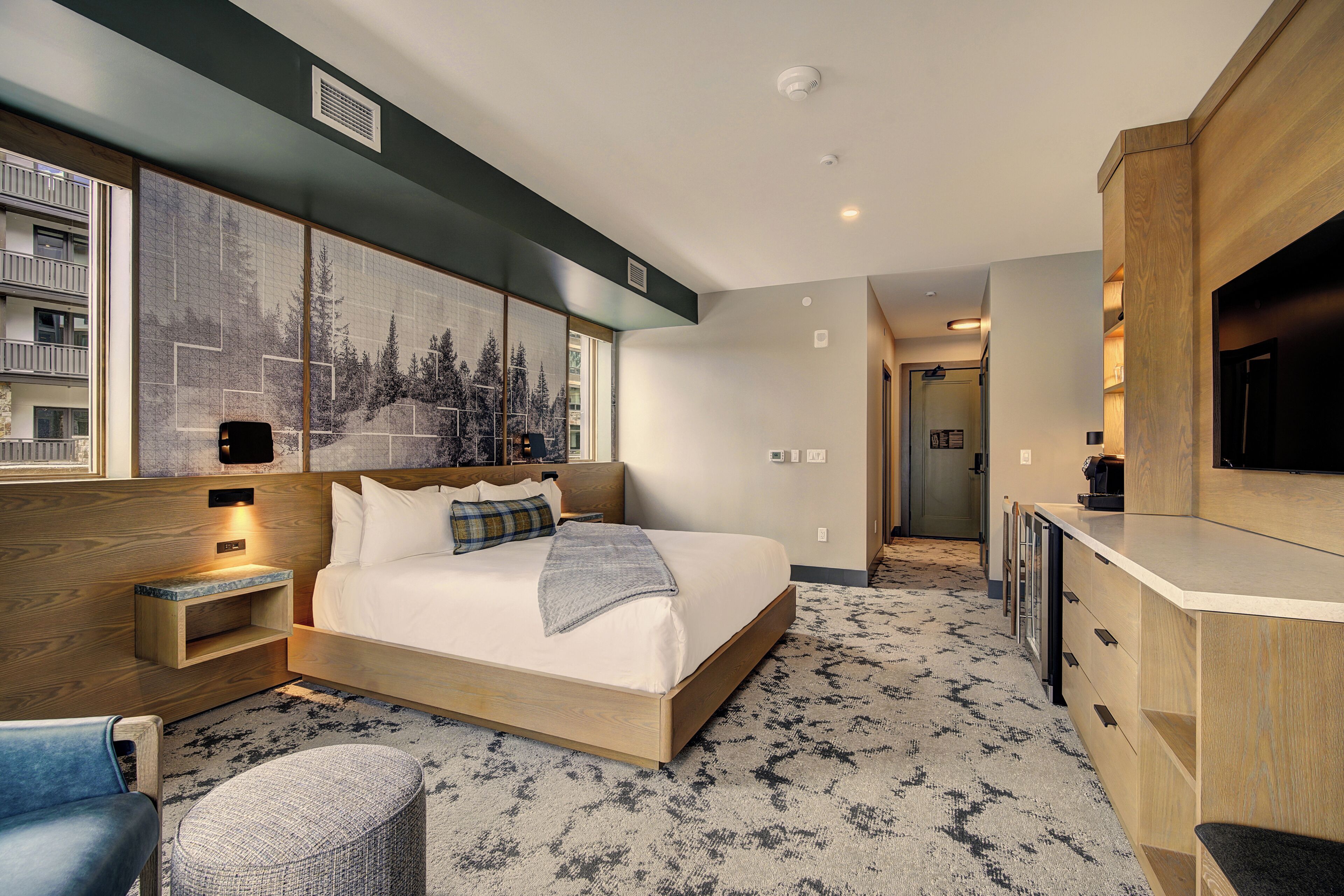 Une chambre d’hôtel équipée d’un lit, d’un bureau, d’une chaise, d’une télévision et d’une grande fenêtre donnant sur la verdure.