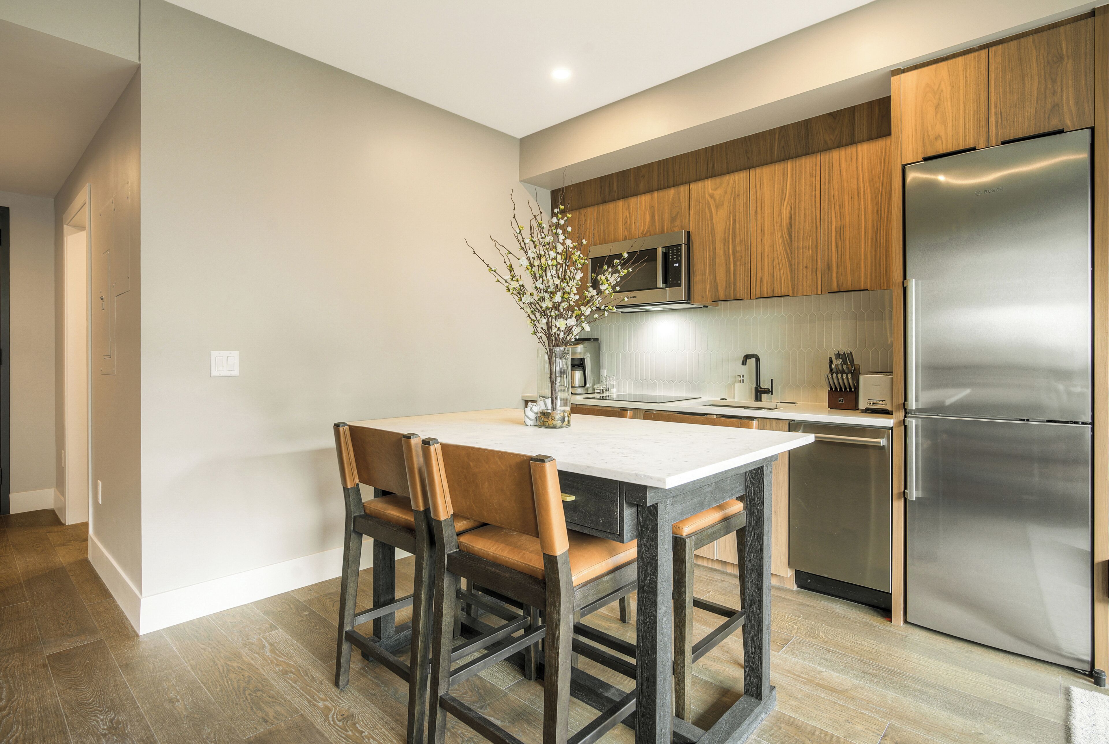 A modern kitchen with a stainless steel refrigerator, wooden cabinets, a marble countertop, and a dining area with a table and chairs.