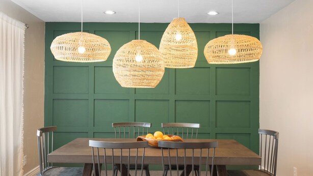 Un coin repas avec un mur lambré vert, une table en bois et quatre suspensions en osier.