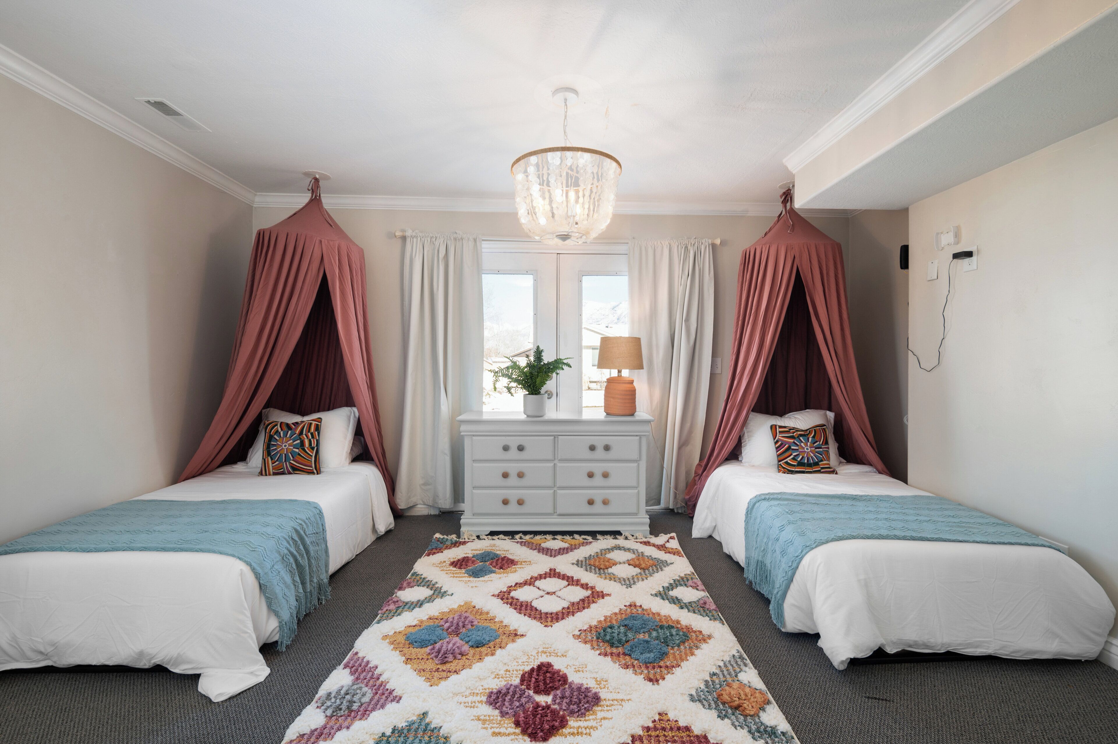 A bedroom with two beds, a chandelier, a dresser, and a window with curtains.