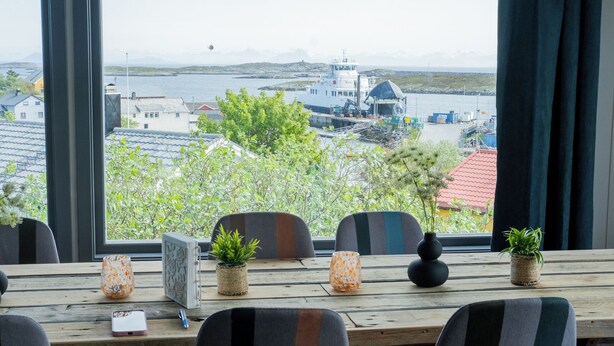 A wooden table with chairs, potted plants, and a view of a coastal town with buildings and a lighthouse. - Træna Overnatting (Traena)