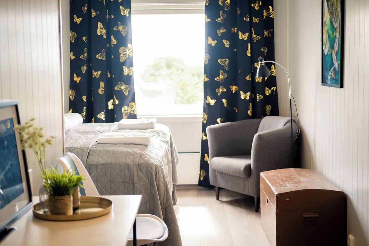 A hotel room with a bed, a chair, a desk with a plant, and a suitcase.