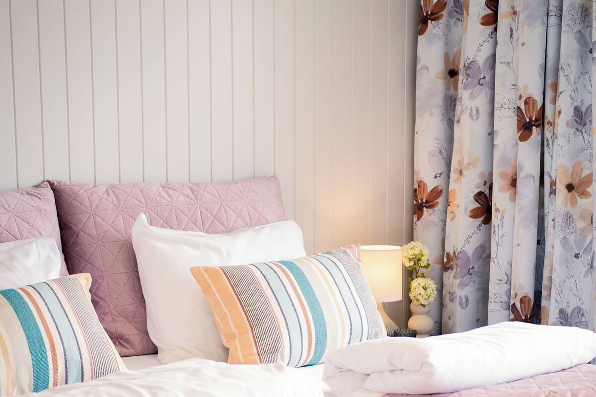 A neatly made bed with a pink quilt, multiple pillows, and a floral curtain.