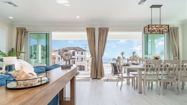 A spacious living area with a wooden coffee table, blue sofa, and a dining set. A large decorative shell is on the table, and the room has large windows with a view of buildings.