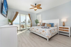 Relax in this serene bedroom with a large bed, nightstand, and TV, offering a tranquil view of palm trees and the ocean.