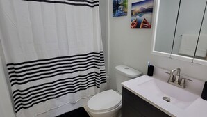 Un baño con inodoro blanco, cortina de ducha a rayas negras y blancas, lavabo con espejo y dos cuadros en la pared.