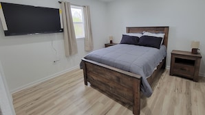Un dormitorio con una cama de madera, un televisor de pantalla plana montado en la pared, un ventanal con cortinas y una mesita de noche con lámpara.