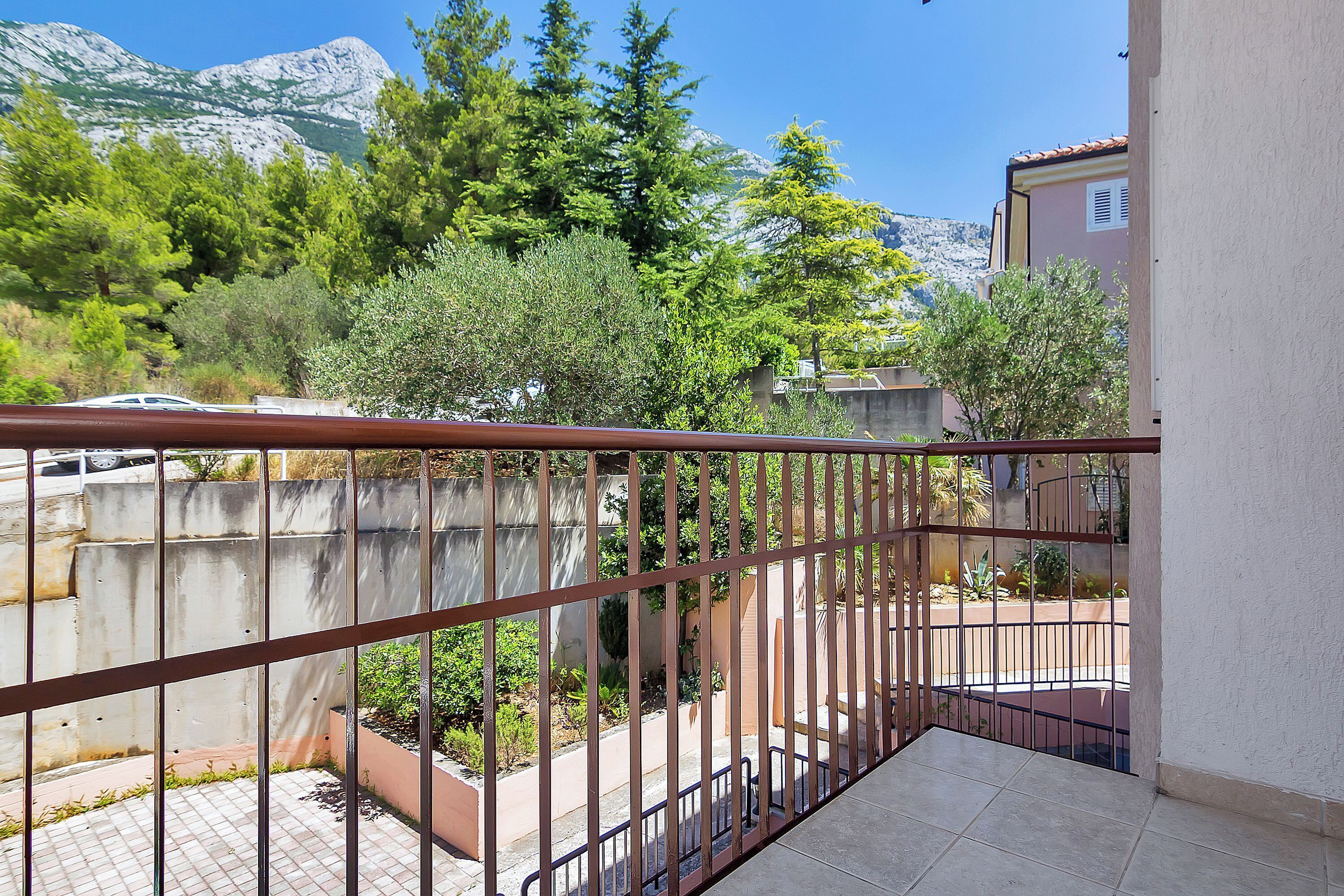 A balcony with a metal railing overlooking a courtyard with trees and a mountain in the background.