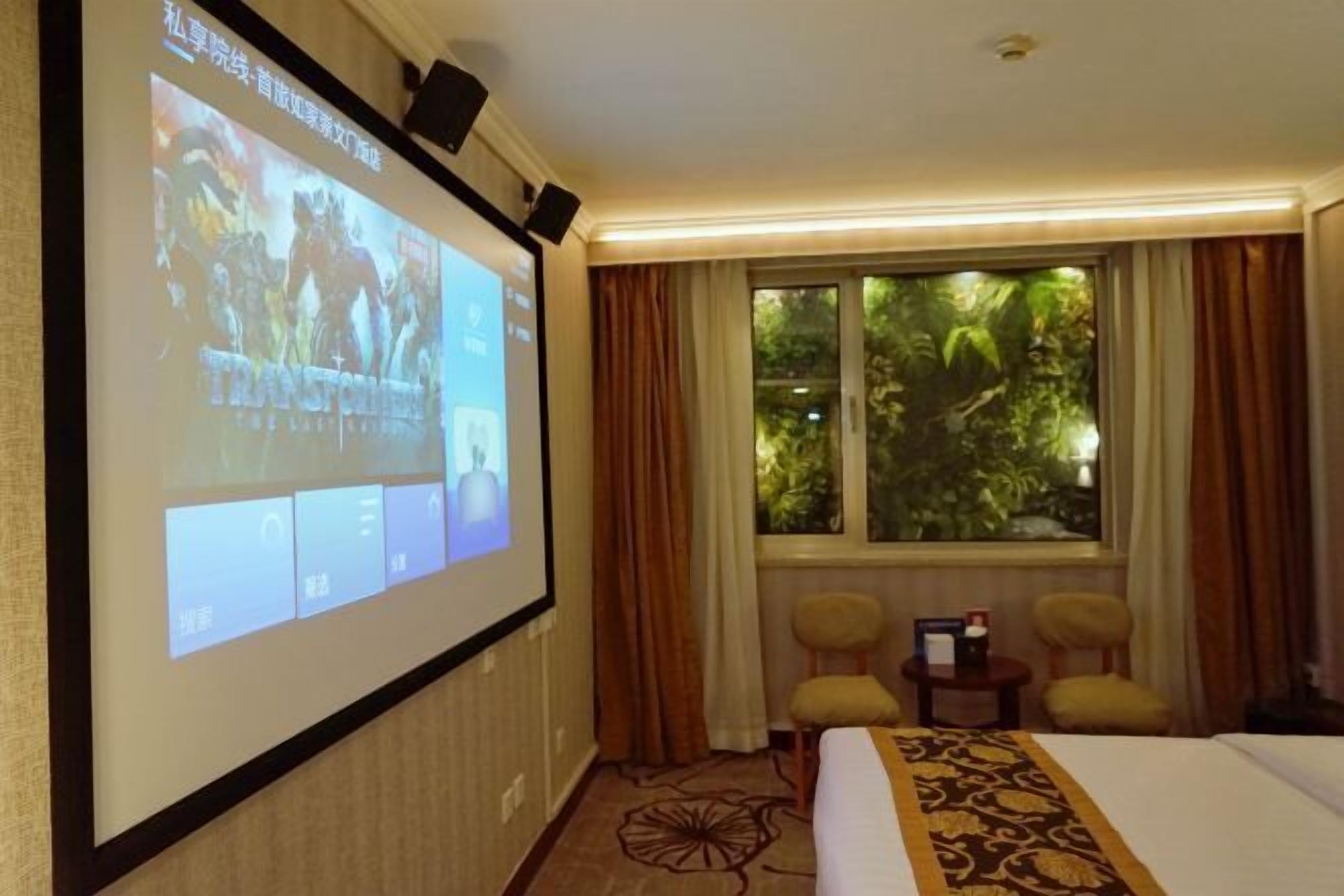 A hotel room with a large screen displaying a movie, a bed, two chairs, a window with a view of greenery, and a carpet with a floral pattern.