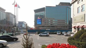 A parking lot with several cars, a building with a digital display, and a flagpole with flags. - Chongwenmen Hotel（Near metro,Tian’anmen Square,Wangfujing,Forbidden city And Temple of heaven） (Beijing)