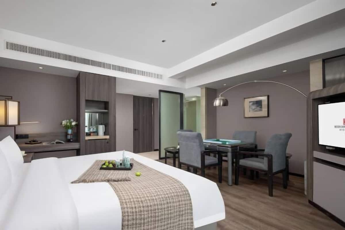 A modern hotel room with a bed, a desk, a dining area, and a TV.
