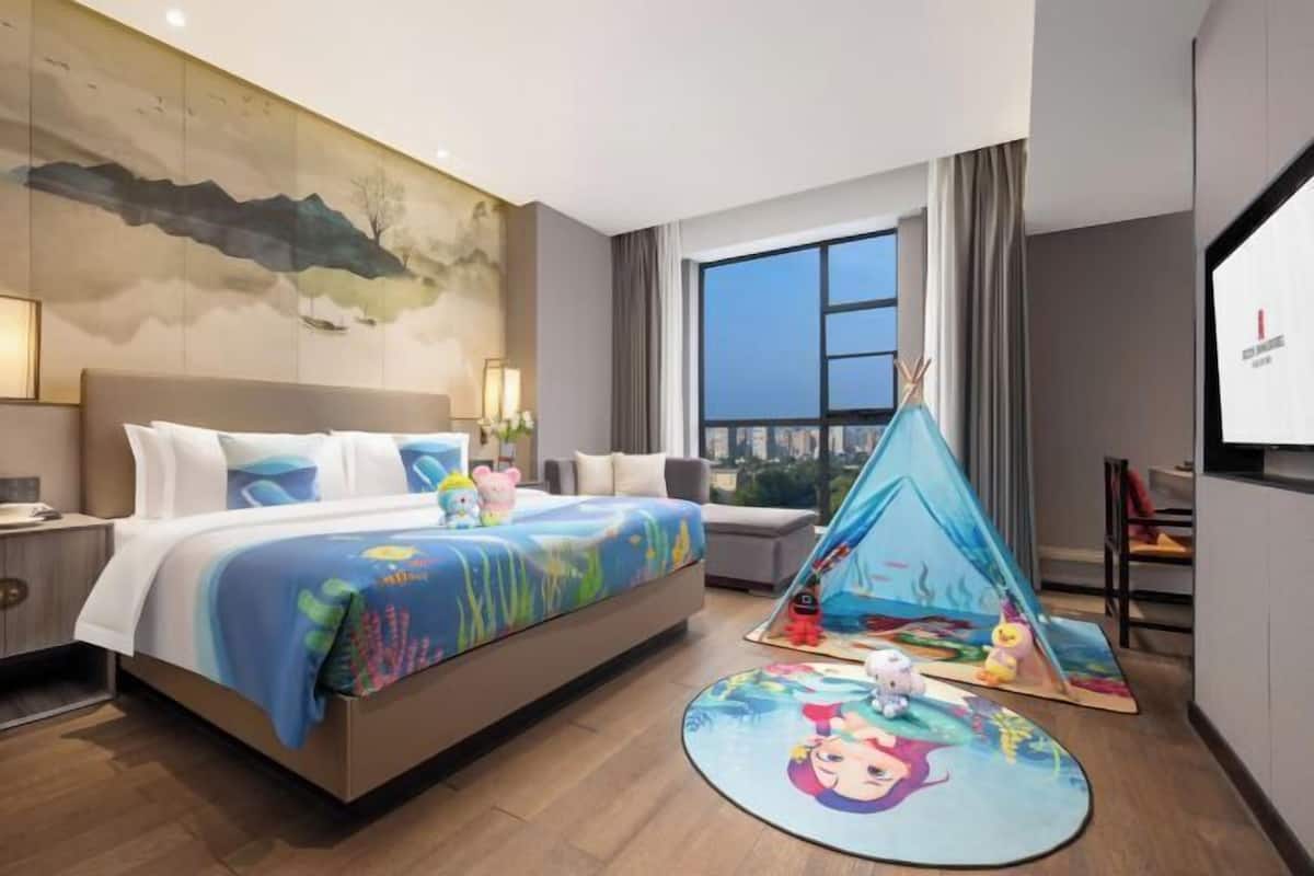 A hotel room with a bed, a large window, a painting on the wall, and a tent set up in the corner.