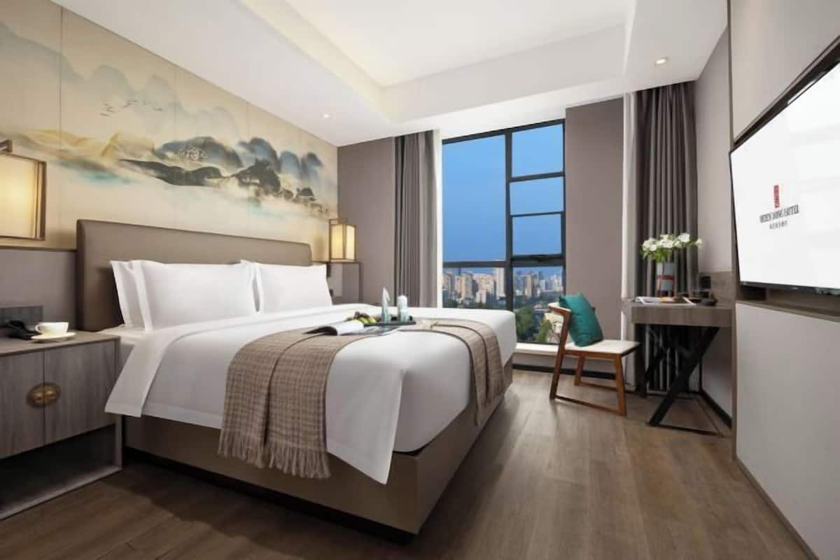 A modern hotel room with a large bed, a desk, a chair, a painting on the wall, and a view of the city.
