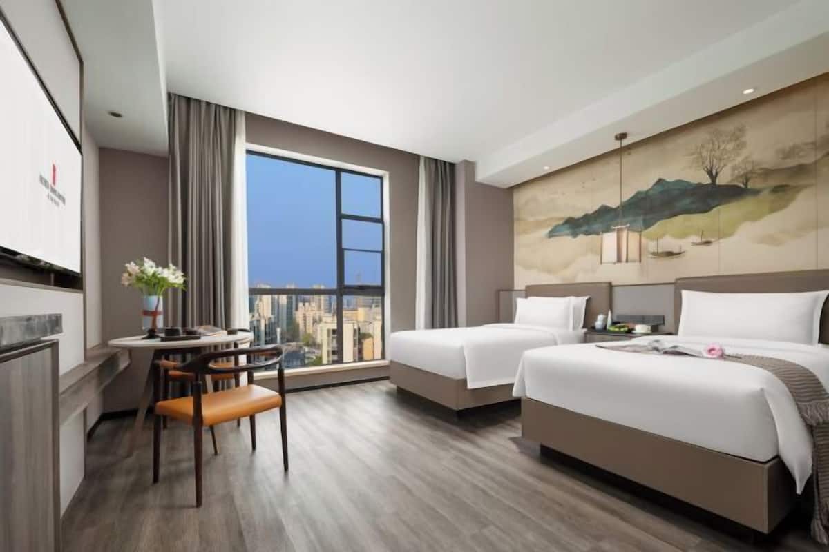 A hotel room with two beds, a dining area with a table and chairs, a large window with a city view, and a painting on the wall.