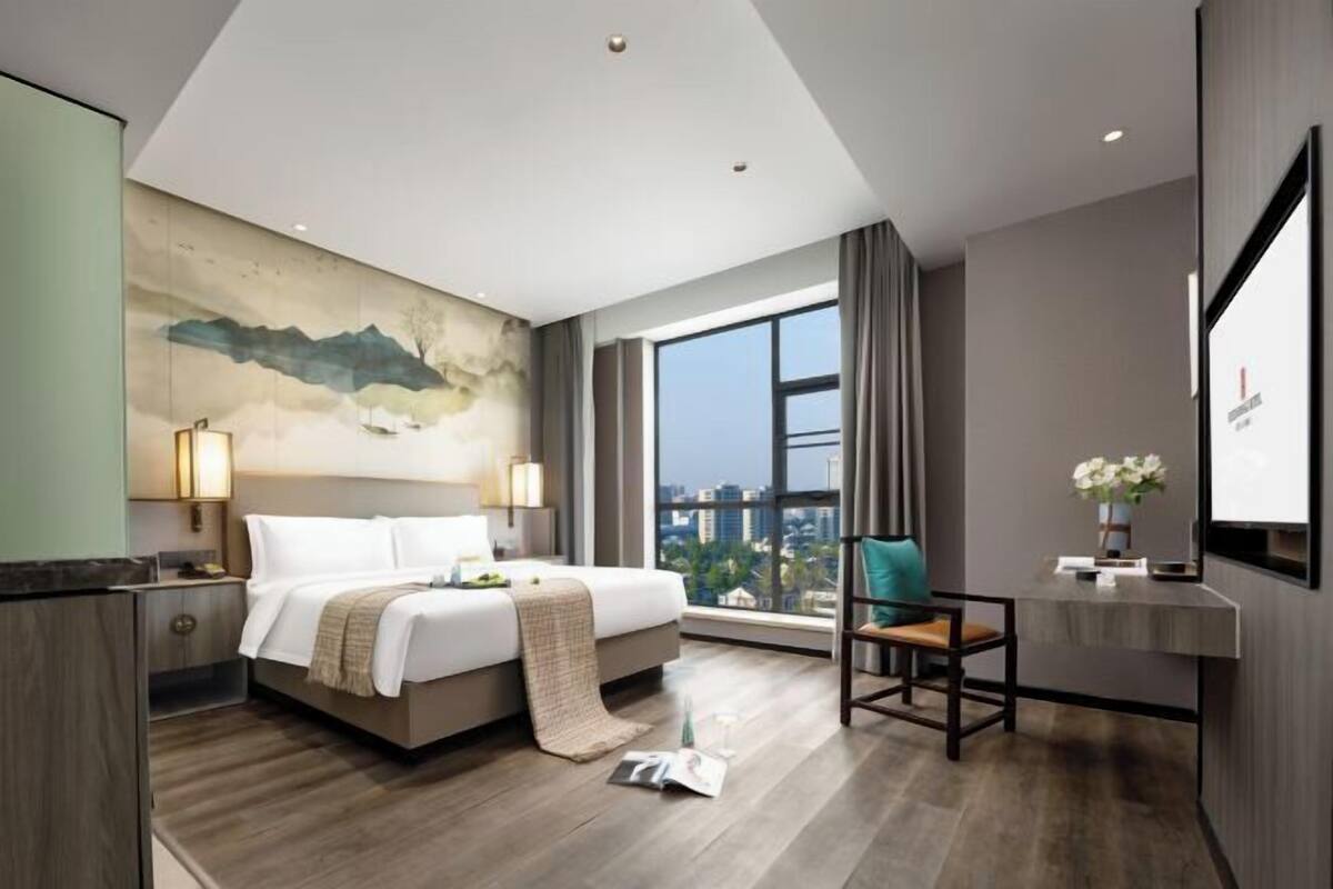 A modern hotel room with a large bed, a desk, a chair, a large window with a city view, and a painting on the wall.