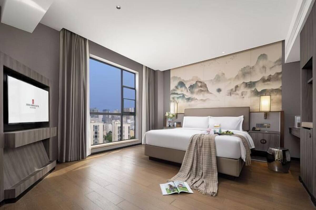 A hotel room with a large bed, a TV, a painting on the wall, and a view of the city.