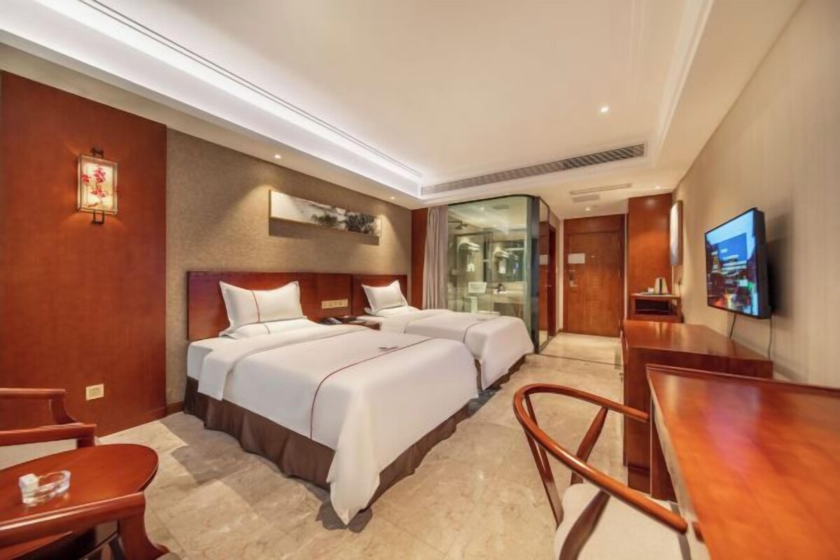 A hotel room with two beds, a desk, a chair, a television, and a bathroom visible through a glass door.