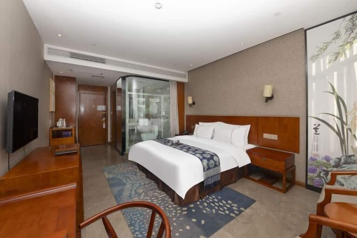 A hotel room with a bed, a desk, a TV, a glass partition, and a painting on the wall.