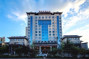 Ein mehrstöckiges Hotel mit einem zentralen Eingang, umgeben von Bäumen und anderen Gebäuden.