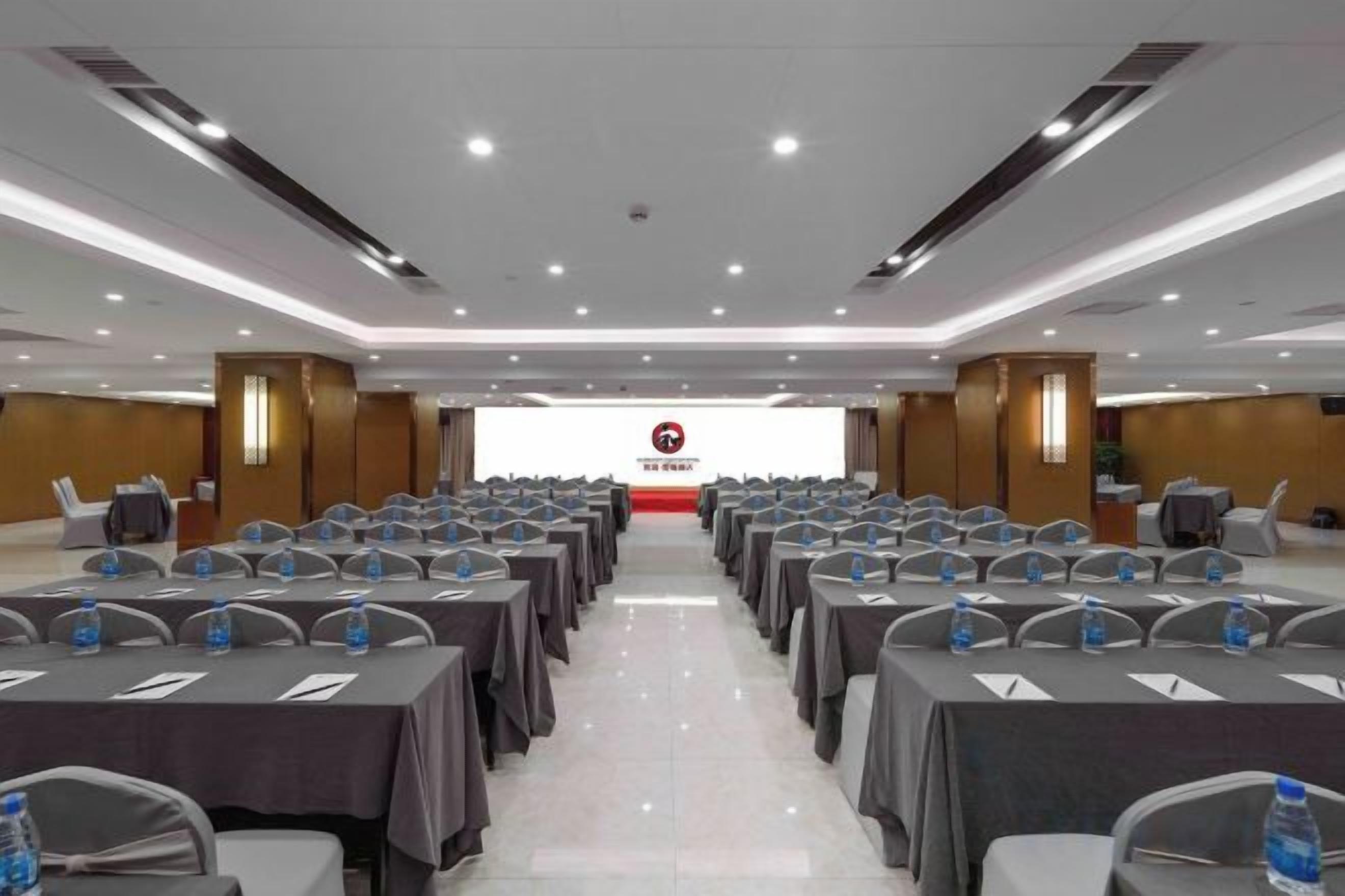 A conference room with rows of tables and chairs, a podium at the front, and a logo on the wall.